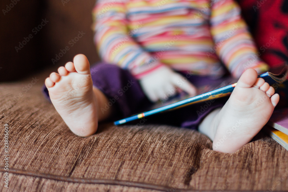 Toddler girl sitting on brown sofa reading a book with focus on her