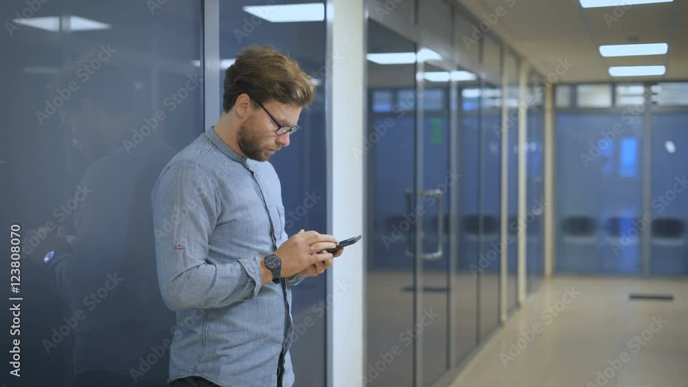 Serious man standing at the wall in an office and writing sms on mobile phone. Serious man standing at the wall in an office and writing sms on mobile phone.