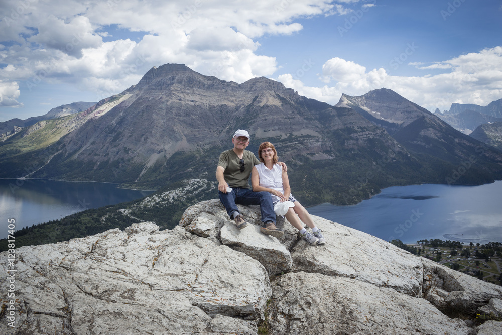 horizontal image of a caucasian husband and wife sitting together on top of a mountain with the lake and mountains in the background - obrazy, fototapety, plakaty 
