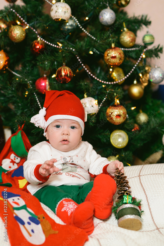 Christmas baby in hat holding red ball near gift box and new year fir tree
