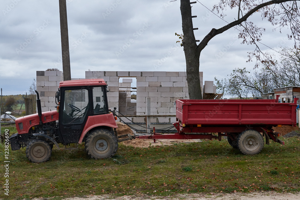  small red tractor with a big trailer in the background of a house under construction in the village on a gloomy autumn day