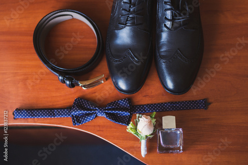 set of groom bow tie, boutonniere, belt, perfume and shoes lie on wooden table