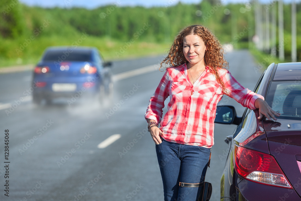 Young woman in red plaid shirt standing near own car on the highway, full length