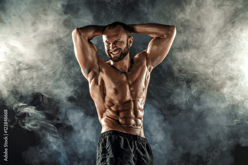 фотографія Studio portrait of topless muscular sportsman posing with arms on head and forced smile over smoke black background
