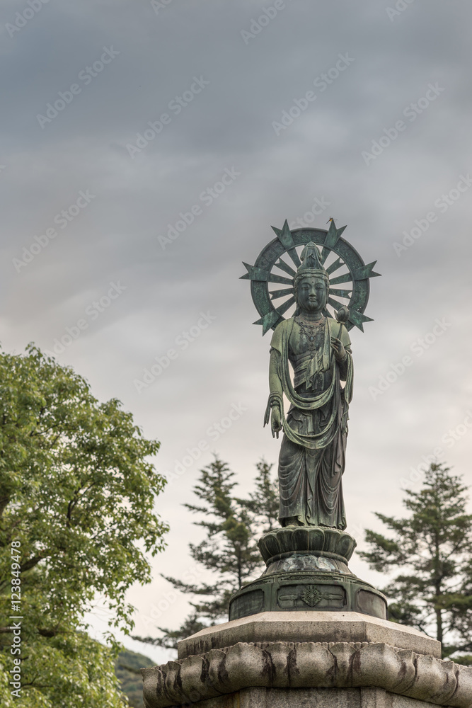 Kyoto, Japan - September 16, 2016: Quan Yin standing in lotus and with halo statue on pedestal in Yuzen-en garden of Chion-in Buddhist Temple. Cloudy skies, some green trees.