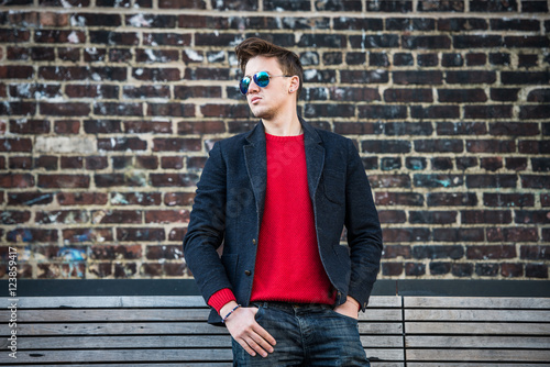 Fashionable young adult stylish man posing outdoors wearing red pulower, jeans, cotton jacket and sunglasses. Stylish handsome caucasian man with messy hairstyle.