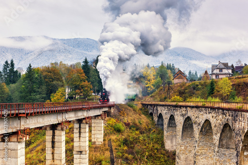 Old steam train passing over railway viaduct in Carpathian mountains, colorful countryside landscape