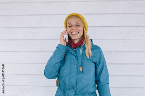 smiling young woman talking on the phone. woman in yellow knitted hat and a blue jacket using smart phone on white wooden background