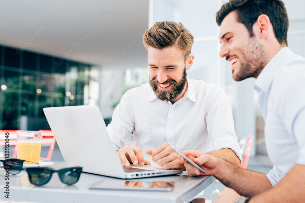 Two young bearded caucasian modern business man sitting in a bar, using laptop, looking downward, tapping on keyboard  - business, work, technology concept