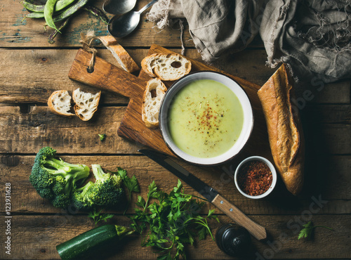 Homemade pea, broccoli, zucchini cream soup in white bowl with fresh baguette on wooden board over rustic background, top view, horizontal composition photo