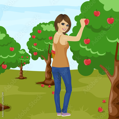 full length portrait of young brunette woman picking red apple from tree in orchard