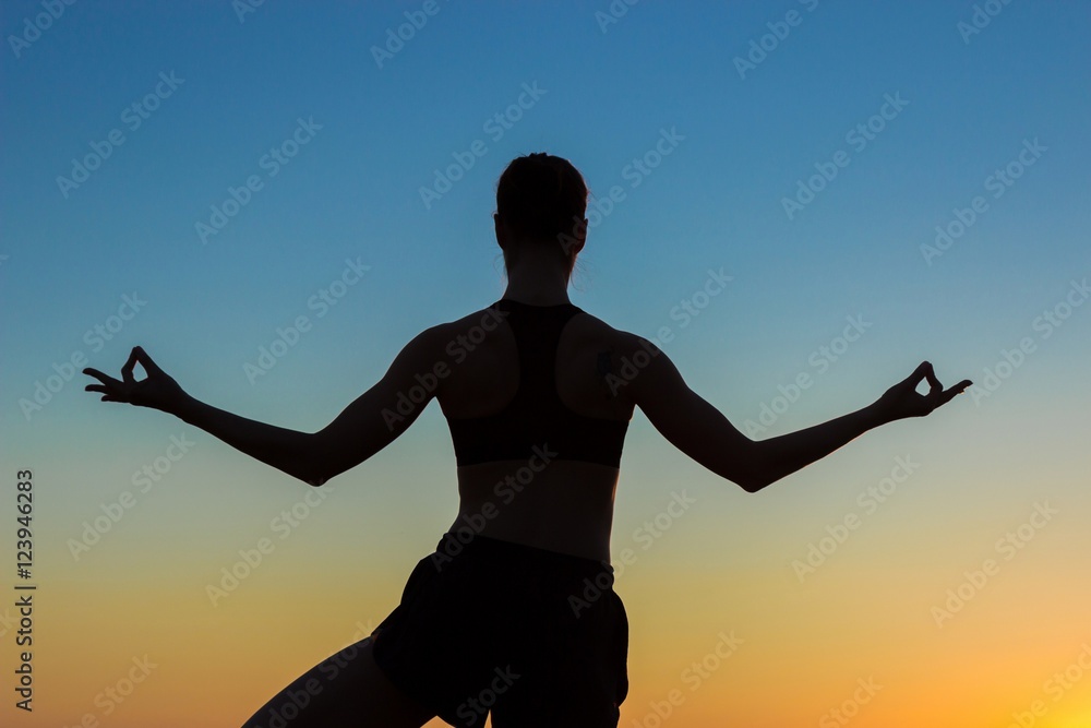 Fototapeta premium Silhouette of sporty woman practicing yoga in the park at sunset - half lotus tree pose. Sunset light, golden hour. Freedom, health and yoga concept