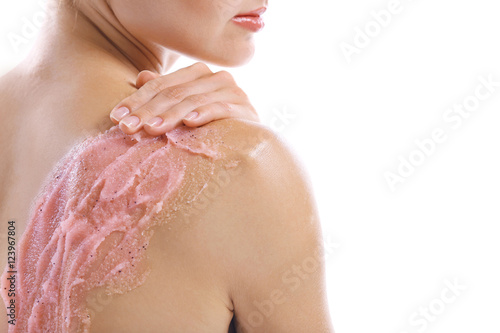 Young woman applying scrub on shoulder on white background