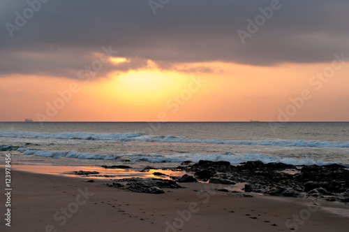The sun rising into a bank of cloud  with rocks and beach in the foreground and ships on the horizon.