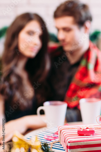 Christmas present, happy couple blurred background. Focus on red new year gift, family with hot chocolate cups backdrop. Holiday, party, winter, coziness concept