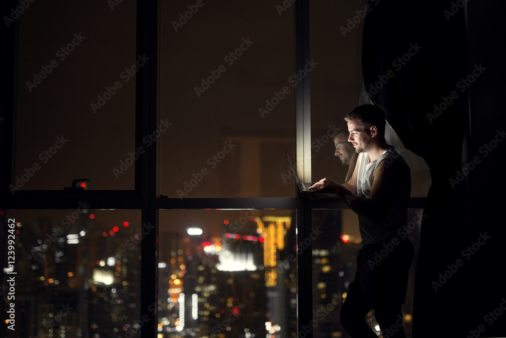 Young designer working on laptop at night near window with beautiful view.