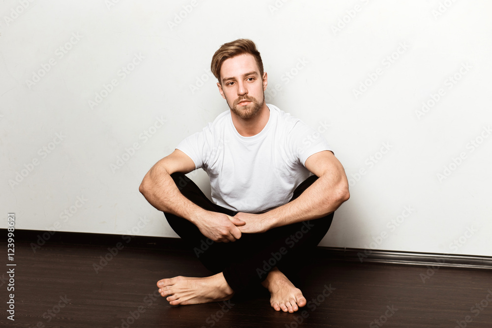 A young caucasian blond man in a white t-shirt and black trousers sitting on the floor A young caucasian blond man in a white t-shirt and black trousers sitting on the floor