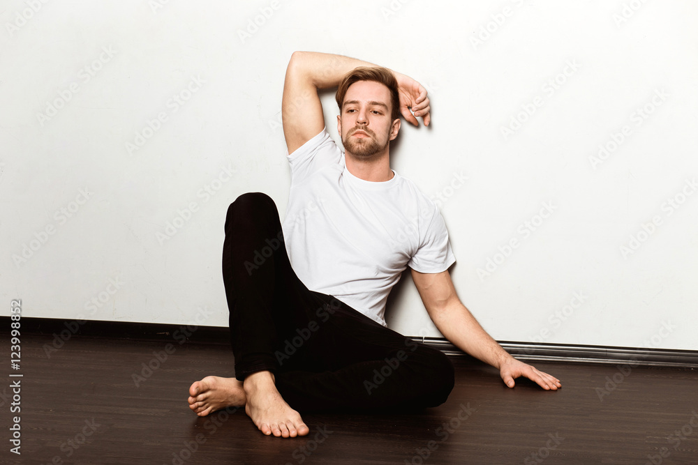 A young caucasian blond man in a white t-shirt and black trousers sitting on the floor