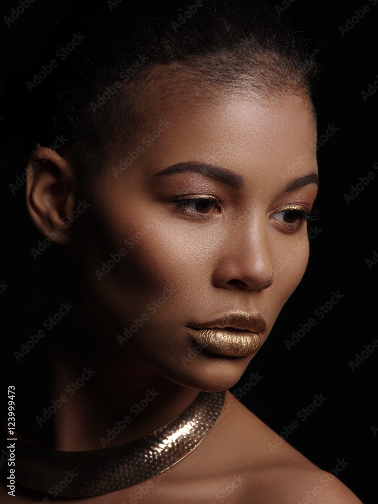 Fototapeta premium Portrait of an extraordinary beautiful naked african american model with perfect smooth glowing skin, make up, full golden lips, shaved haircut and gold jewelry on her neck, dark background
