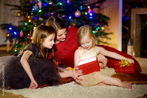 Young mother and her two daughters unwrapping Christmas gifts by a fireplace