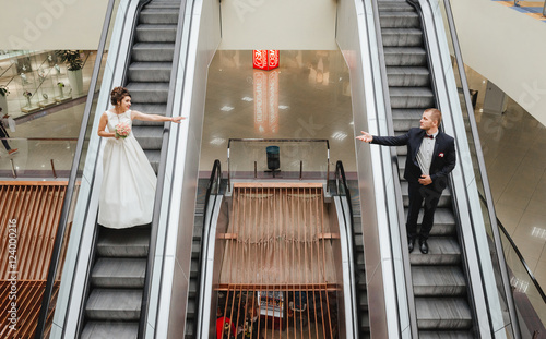 Smiling bride and groom on the rise escalators are located in parallel, drawn to each other hands. View from above