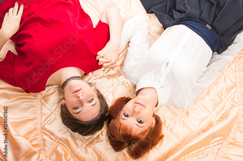 Two lesbian girls lying on the blanket cover and hold hands.