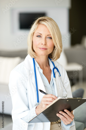 Attractive blond woman doctor nurse practitioner in lab coat writing on clipboard in clinic office hospital