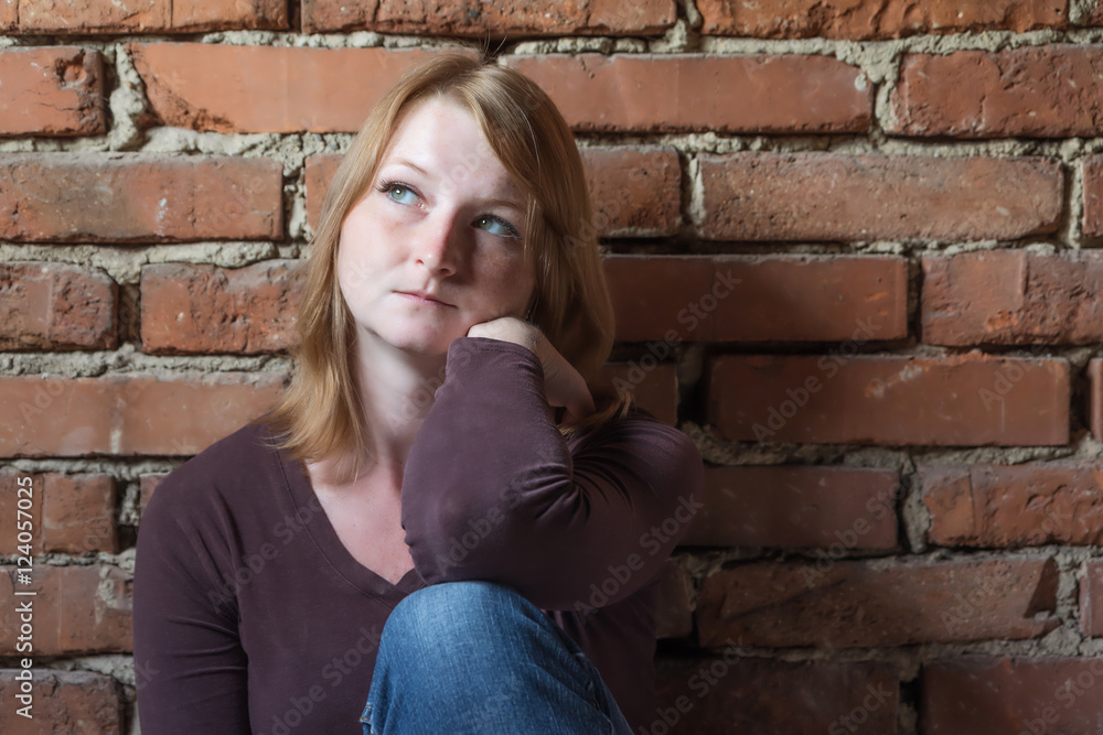 Portrait of serious long haired young woman sitting in front of brick wall. Woman is looking to the natural light. Portrait of serious long haired young woman sitting in front of brick wall. Woman is looking to the natural light.