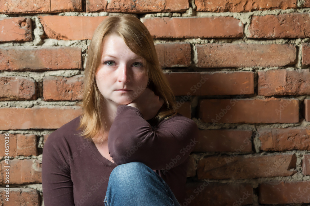 Portrait of pensive young woman sitting in front of brick wall in natural light. Woman is looking at the camera.