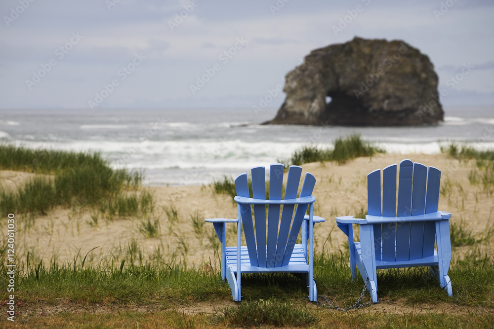 Two Blue Adirondack Chairs On A Grassy Beach With Rock Formations In The Ocean; Rockaway Beach, Oregon, United States of America Two Blue Adirondack Chairs On A Grassy Beach With Rock Formations In The Ocean; Rockaway Beach, Oregon, United States of America