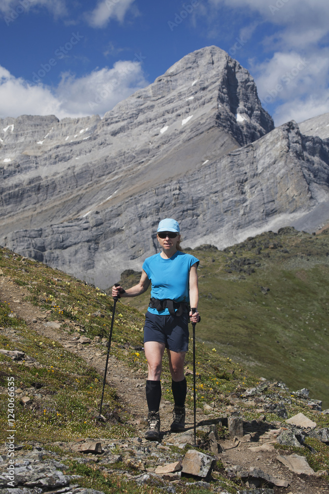 Female Hiker With Hiking Poles On A Mountain Trail With Mountain Peak In The Background And Blue Sky With Clouds; Alberta, Canada Female Hiker With Hiking Poles On A Mountain Trail With Mountain Peak In The Background And Blue Sky With Clouds; Alberta, Canada