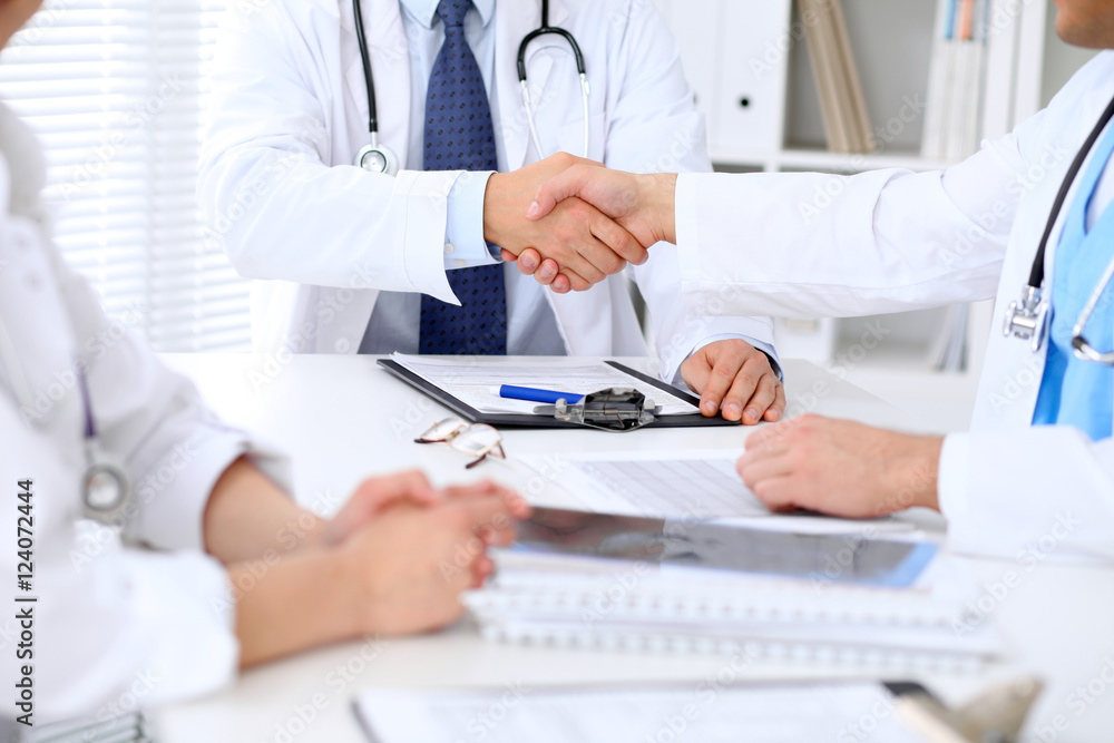 Two doctors shaking hands to each other sitting at the table in hospital office Two doctors shaking hands to each other sitting at the table in hospital office
