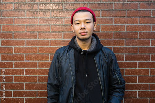 Handsome young asian man.Wearing black jacket and red beanie. looking at camera. Standing in front of brick wall