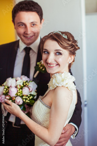 Blue-eyed bride smiles while groom hugs her waist standing in th