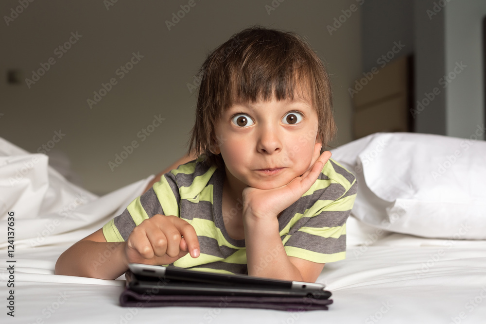 Little boy with digital tablet rests, surprised on the bed on home interior background Little boy with digital tablet rests, surprised on the bed on home interior background