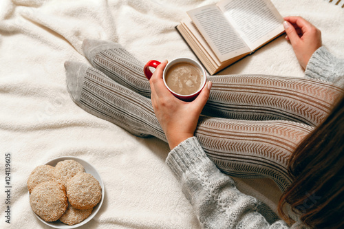 A young woman in a sweater and stockings, with biscuits and red coffee cup in hands relaxing in bed. top view