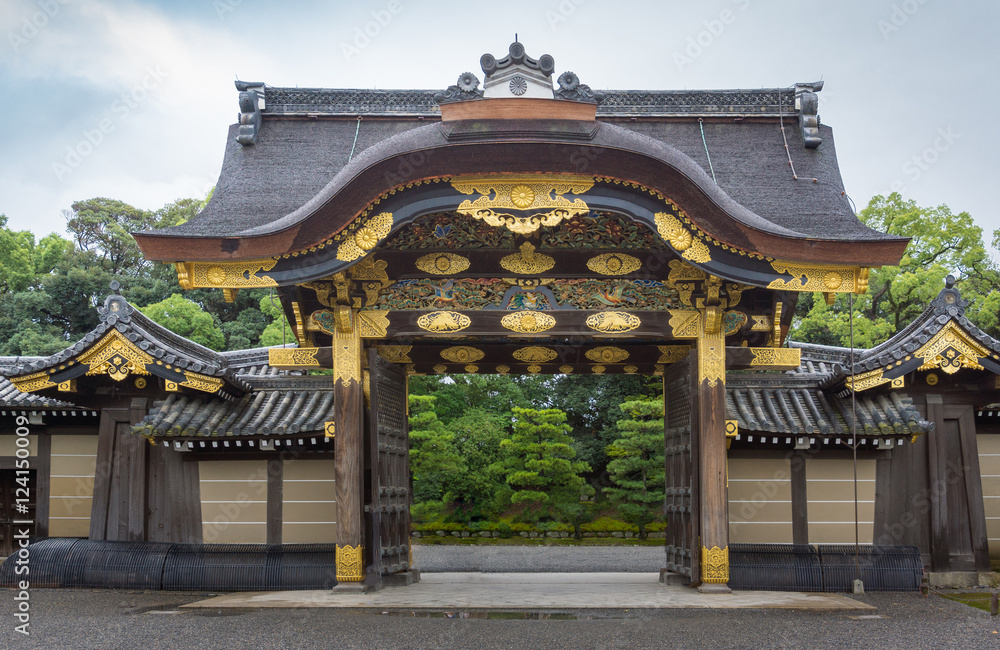 Kyoto, Japan - September 19, 2016: Majestic Kara-mon gate at the Nijo Castle. Plenty of gold trimmings and colorful woodwork. Cloudy sky and green foliage in background.