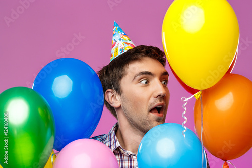 Surprised man celebrating birthday, holding colorful baloons over purple background. © Cookie Studio