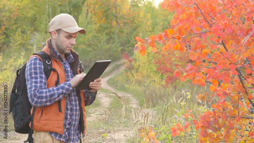 Young, Lost Man With Tablet Looking For Direction On The Forest. Caucasian male outdoors in nature. Hiker young man with backpack in autumn forest. Young, Lost Man With Tablet Looking For Direction On The Forest. Caucasian male outdoors in nature. Hiker young man with backpack in autumn forest.