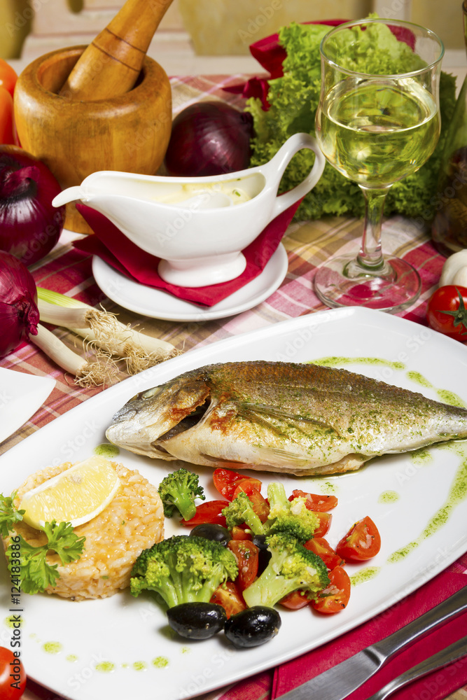 Fried sea bream on a plate with a side dish of broccoli, cherry tomatoes, rice, olives and lemon served beside sauce and white wine. Fried sea bream on a plate with a side dish of broccoli, cherry tomatoes, rice, olives and lemon served beside sauce and white wine.