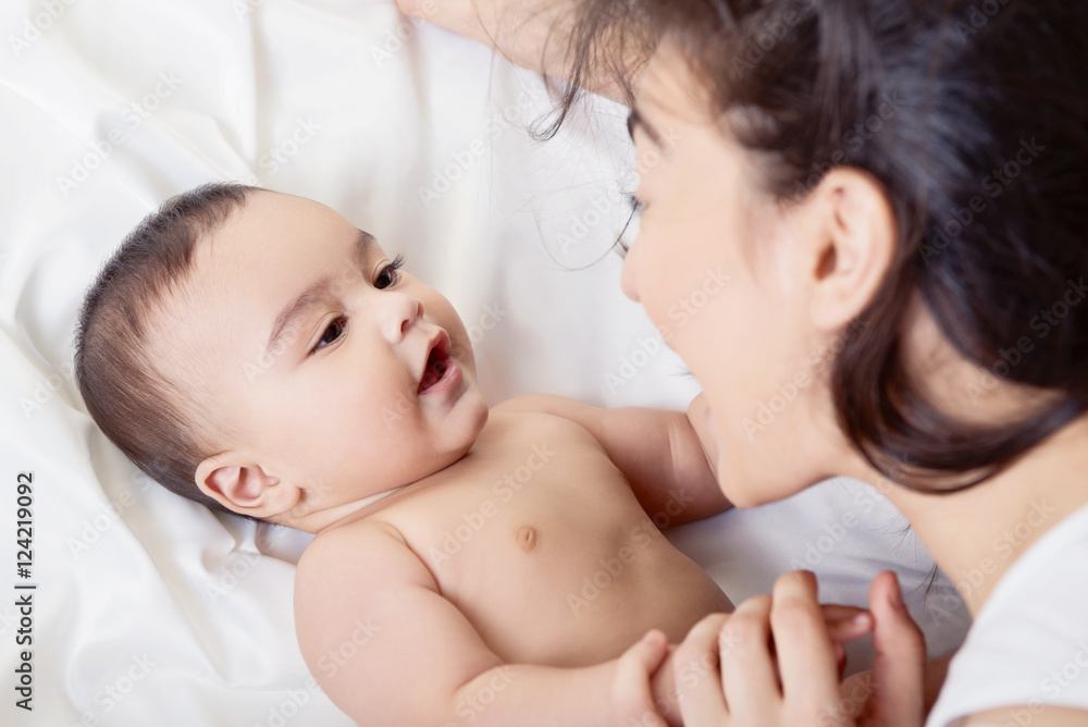 Young mother playing with her baby on white silk bed sheet. Cute Young mother playing with her baby on white silk bed sheet. Cute