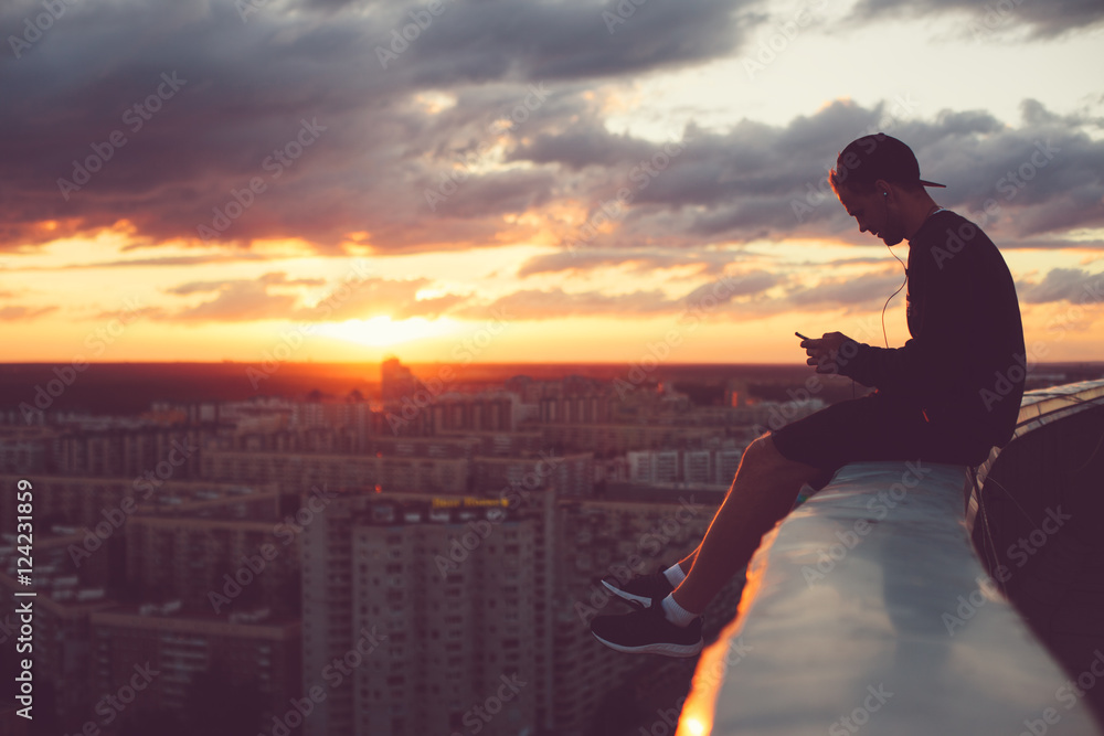 Young brave man sitting above the city with smartphone at sunset - obrazy, fototapety, plakaty 
