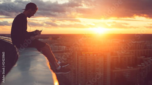 Young risky man chilling above the city with smartphone at sunset