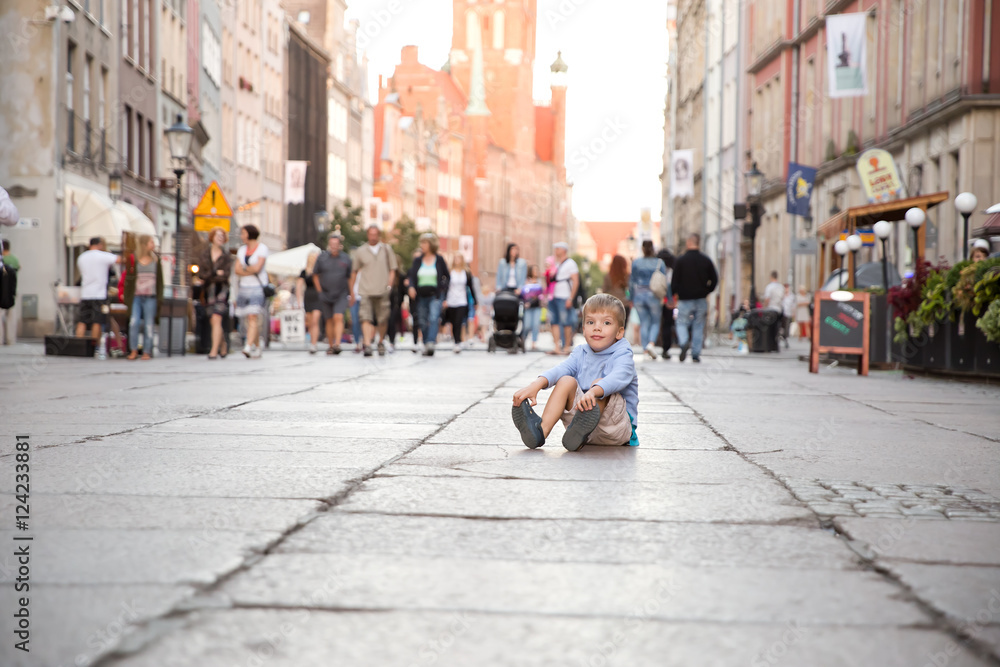 a little blond boy sitting on a ground in a city center