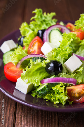 Greek salad (lettuce, tomatoes, feta cheese, cucumbers, black olives, purple onion) on dark wooden background close up. Healthy food.