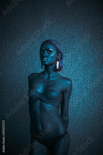Thin woman covered with blue paint poses at the wall