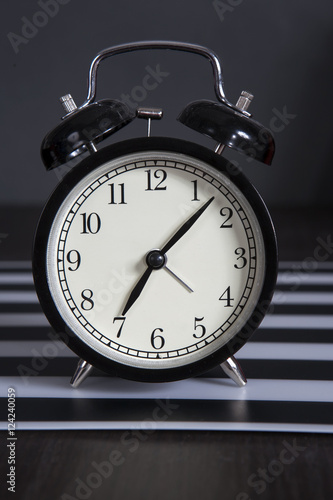 Black alarm clock on a black and white striped napkin showing 7 o'clock on a bedside table