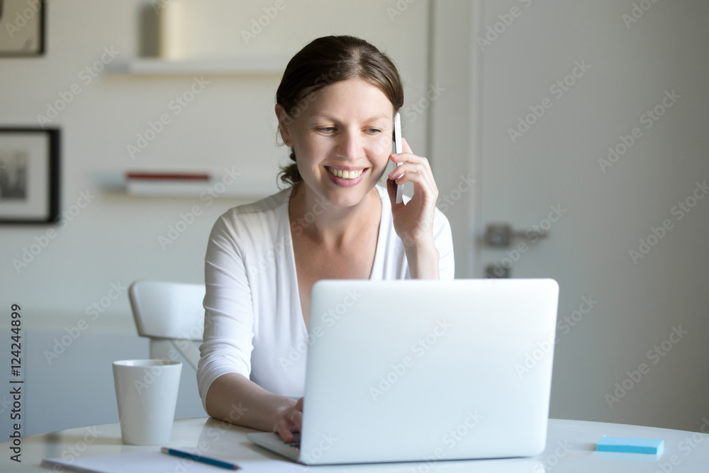 Portrait of a young smiling woman working at the desk with a laptop, looking at the screen, talking on the phone. Business concept photo, lifestyle Portrait of a young smiling woman working at the desk with a laptop, looking at the screen, talking on the phone. Business concept photo, lifestyle