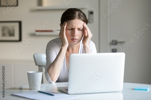 Portrait of a young woman at the desk with a laptop, holding tight her temples. Business concept photo, lifestyle