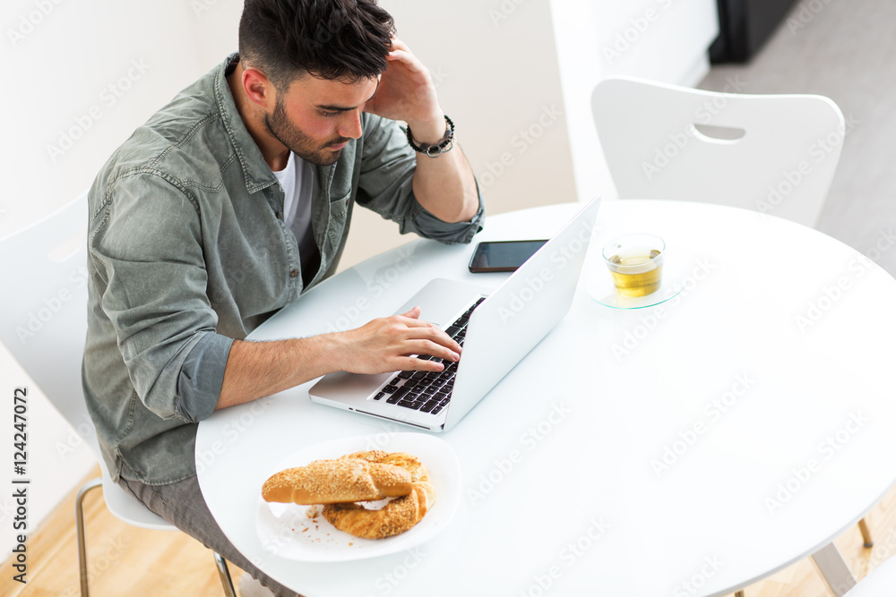 Young man doing something on laptop in his kitchen.Looking worried. Young man doing something on laptop in his kitchen.Looking worried.